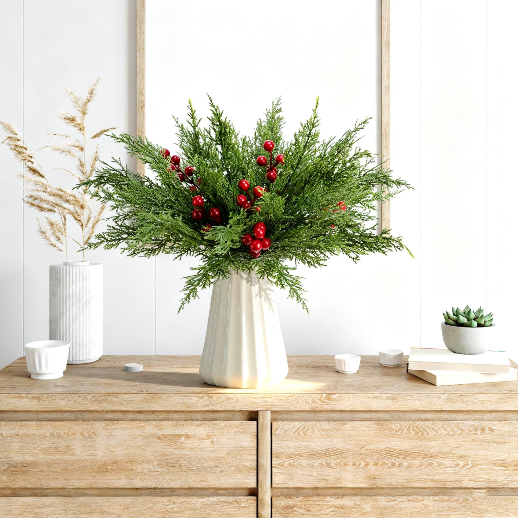 Artificial pine branches with red berries in a vase on a wooden dresser, with decorative items.
