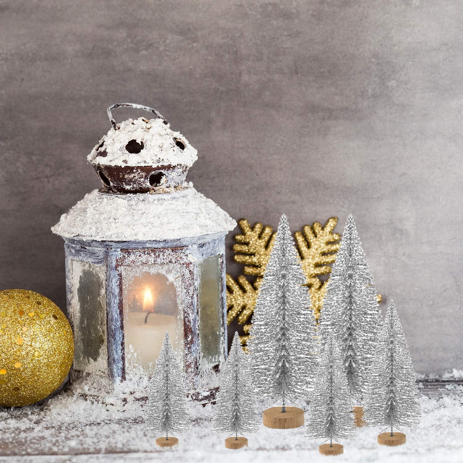 Miniature silver glitter Christmas trees with wooden bases arranged with a lantern, candle, and gold ornaments.