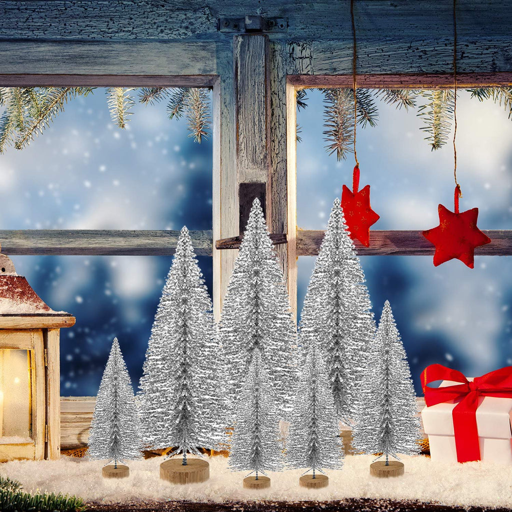 Silver glitter bottle brush Christmas trees arranged on a snowy windowsill with a lantern and gift box.