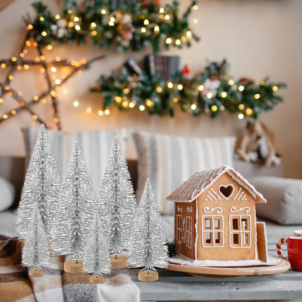 Silver glitter bottle brush Christmas trees and a gingerbread house on a table with festive lights in the background.