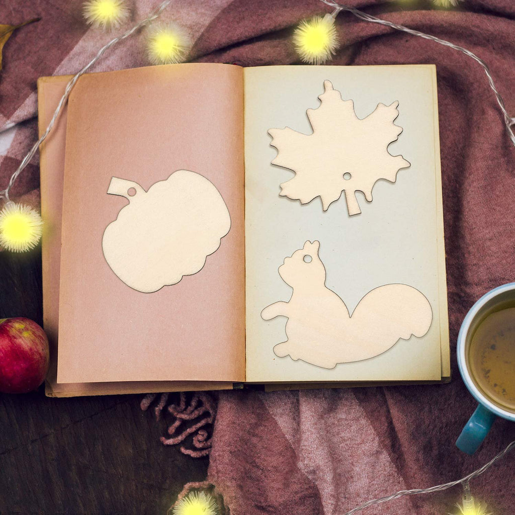 Unfinished wood Thanksgiving ornaments in pumpkin, maple leaf, and squirrel shapes laid open on a book with a cup of tea and apple nearby.