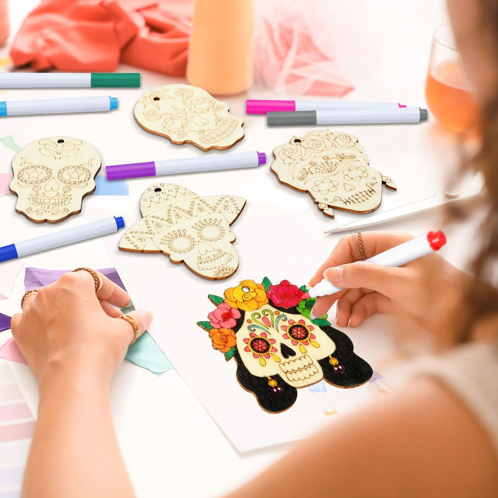 Hands coloring a wooden sugar skull ornament with markers, surrounded by other unfinished ornaments and coloring pens.