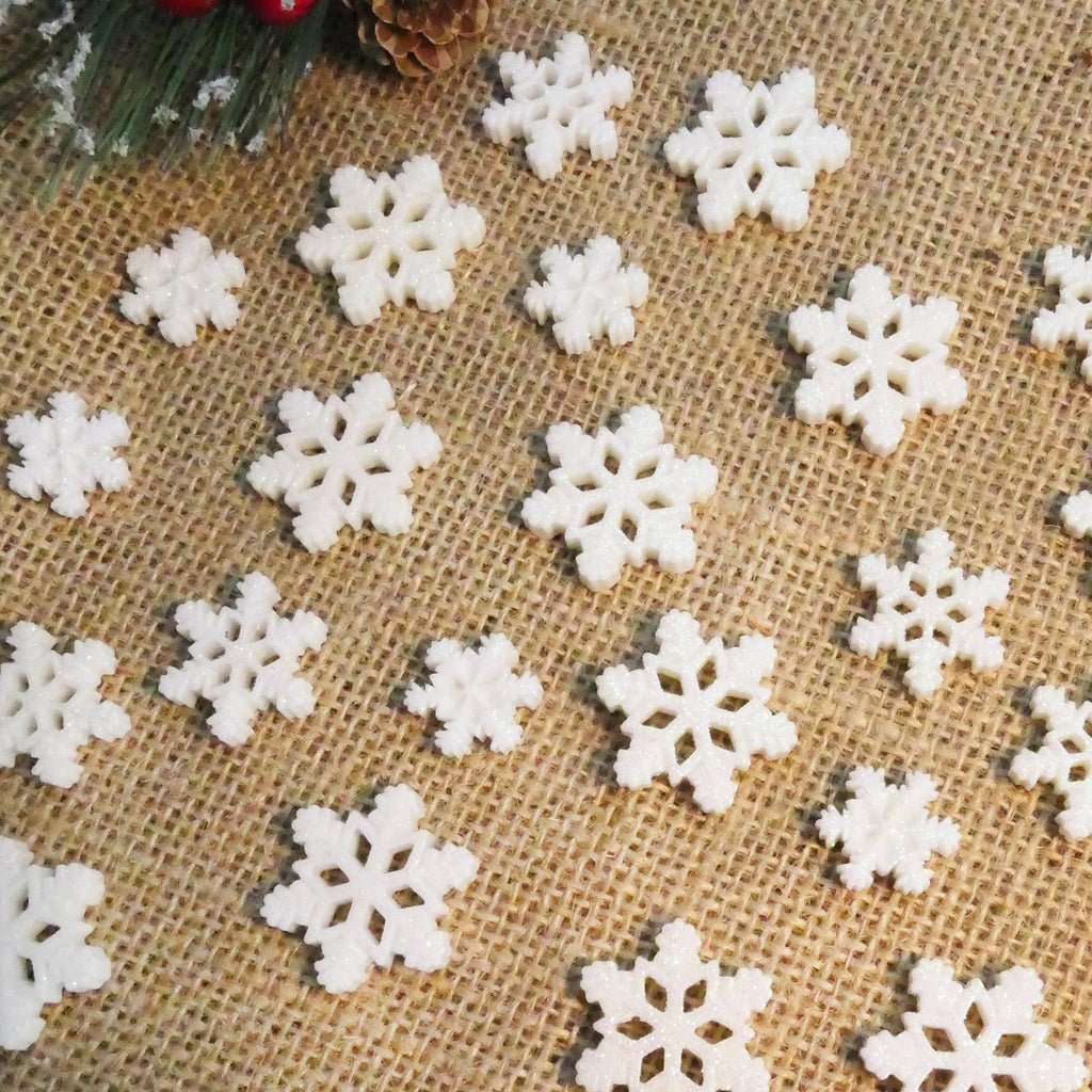 Assortment of white glitter resin snowflakes in three sizes scattered on a burlap background with pinecone and greenery accents.