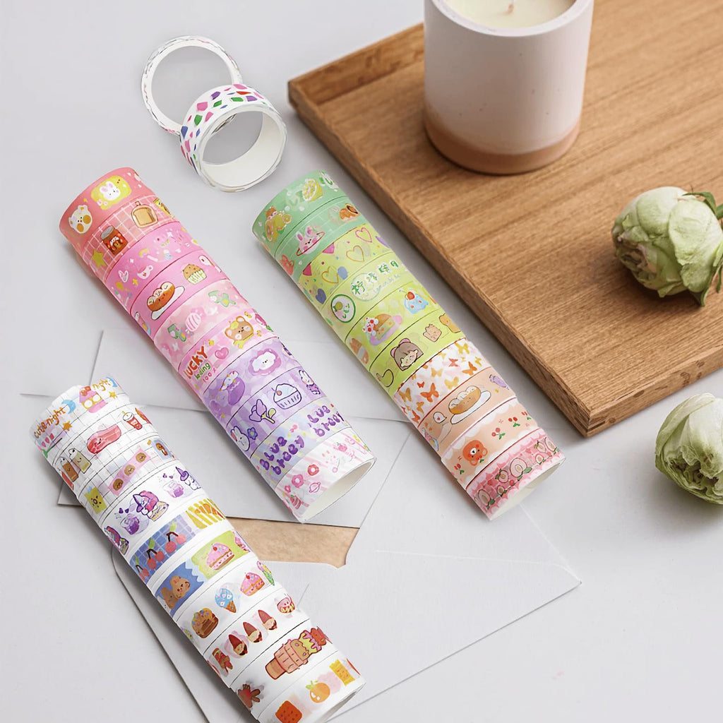 Three rolls of decorative washi tape with colorful patterns, including food and animal designs, arranged on a white surface with a wooden tray and candle in the background.