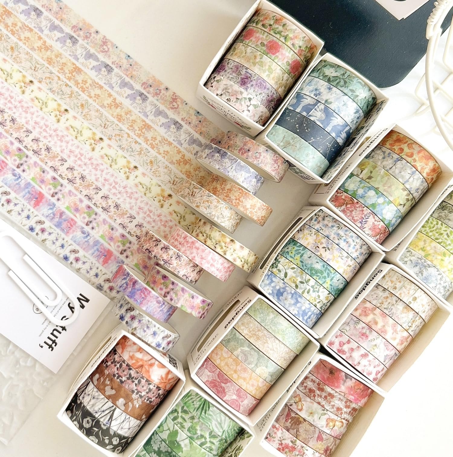 Collection of floral washi tape rolls in various pastel and muted colors, arranged in boxes and fanned out on a white surface.