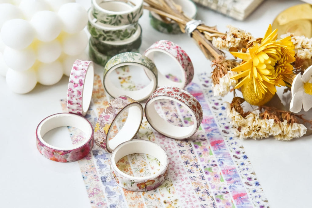 Assortment of floral washi tape rolls and loose tape strips arranged with dried flowers and a white bubble candle.
