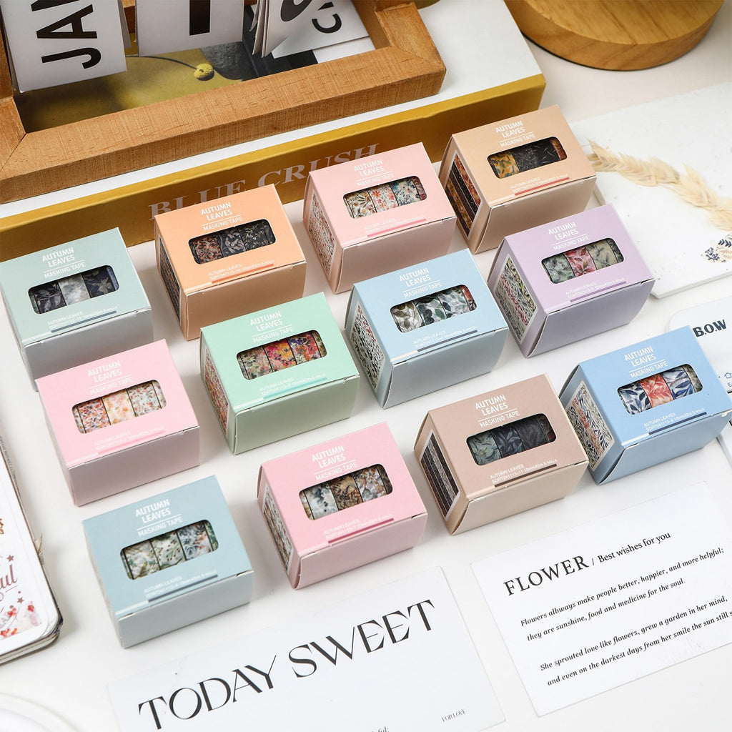 Collection of colorful masking tape boxes with floral and leaf patterns, arranged on a white surface with decorative cards.
