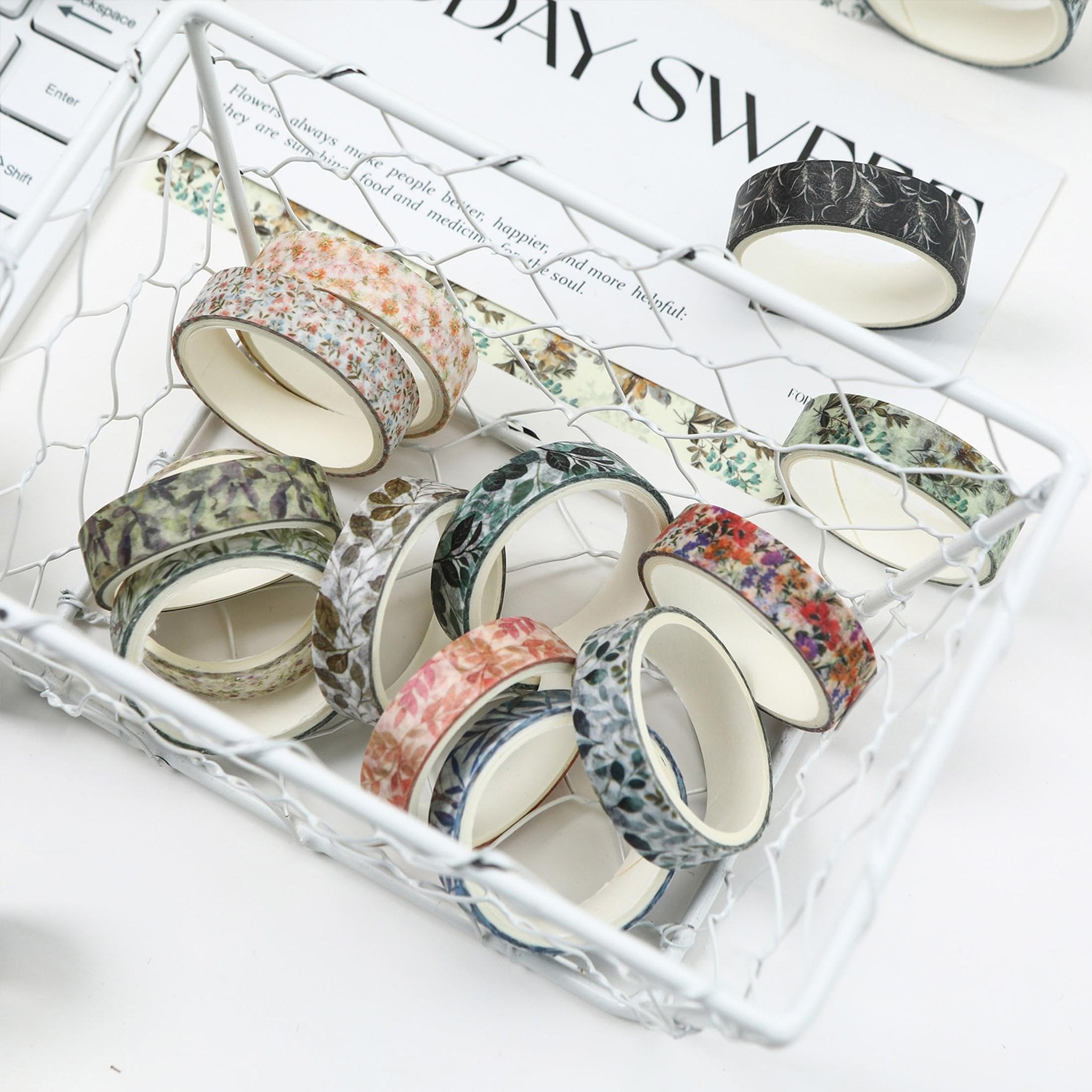 Assortment of floral and leaf pattern washi tape rolls in a white wire basket on a white surface.
