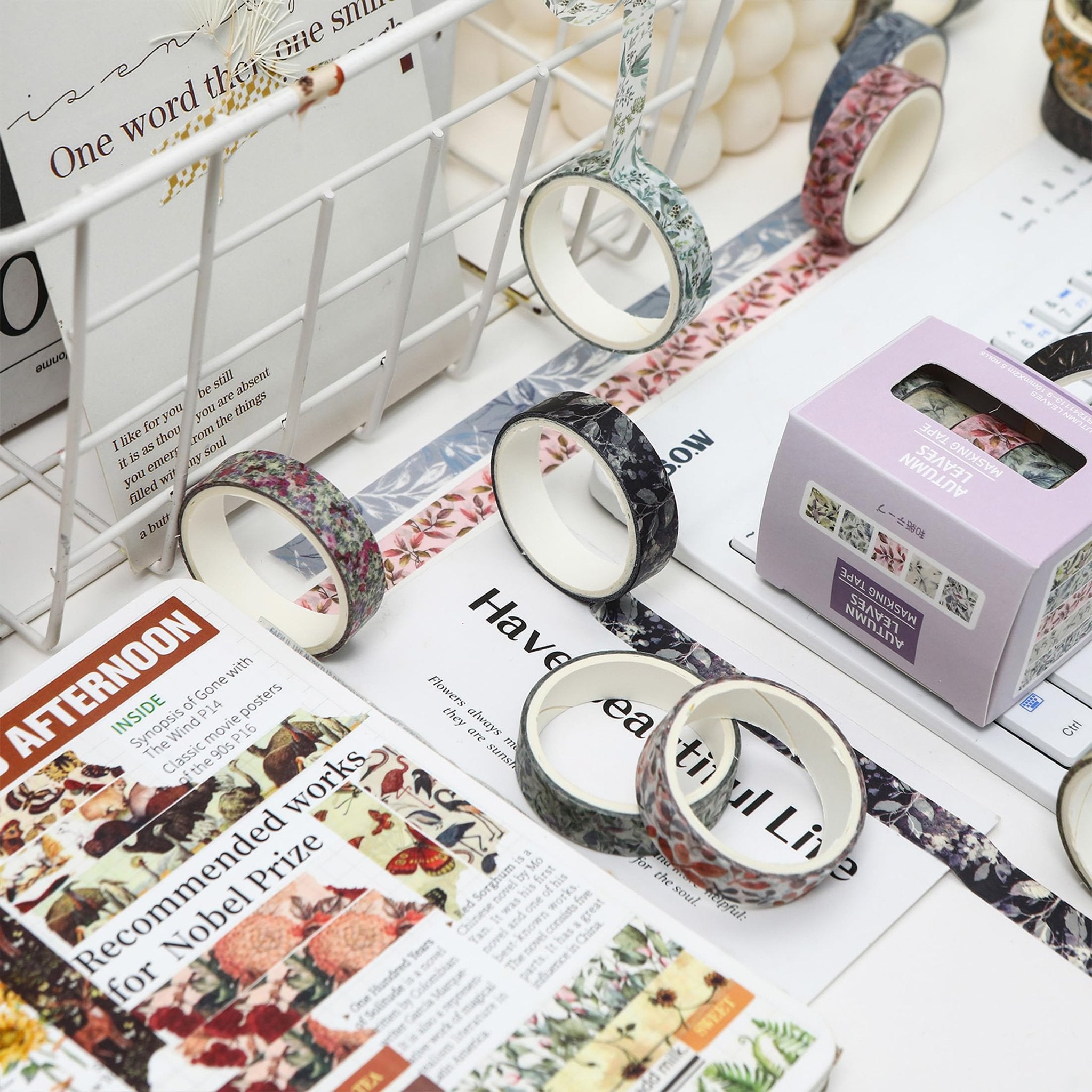 Assortment of floral and leaf patterned washi tape rolls and a box of washi tape on a white surface with scattered papers.