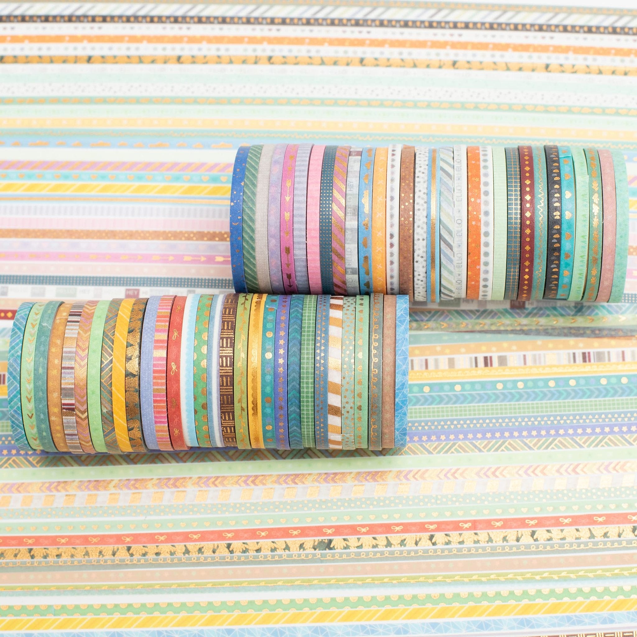 Two stacks of skinny gold foil washi tape rolls with various patterns on a striped background.