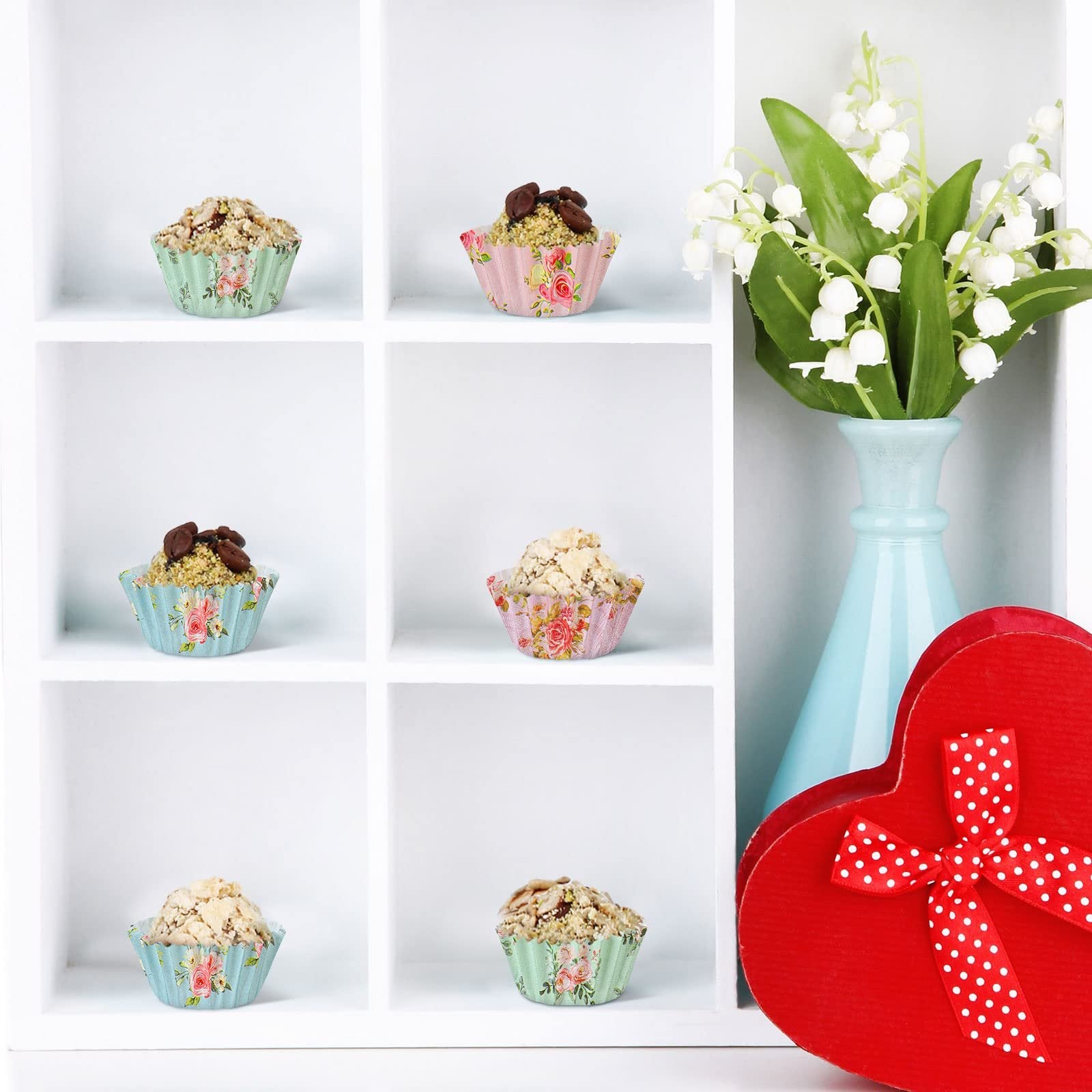 Six floral patterned mini cupcake liners with baked goods displayed in a white cubby shelf, with a vase of flowers and a heart-shaped box.