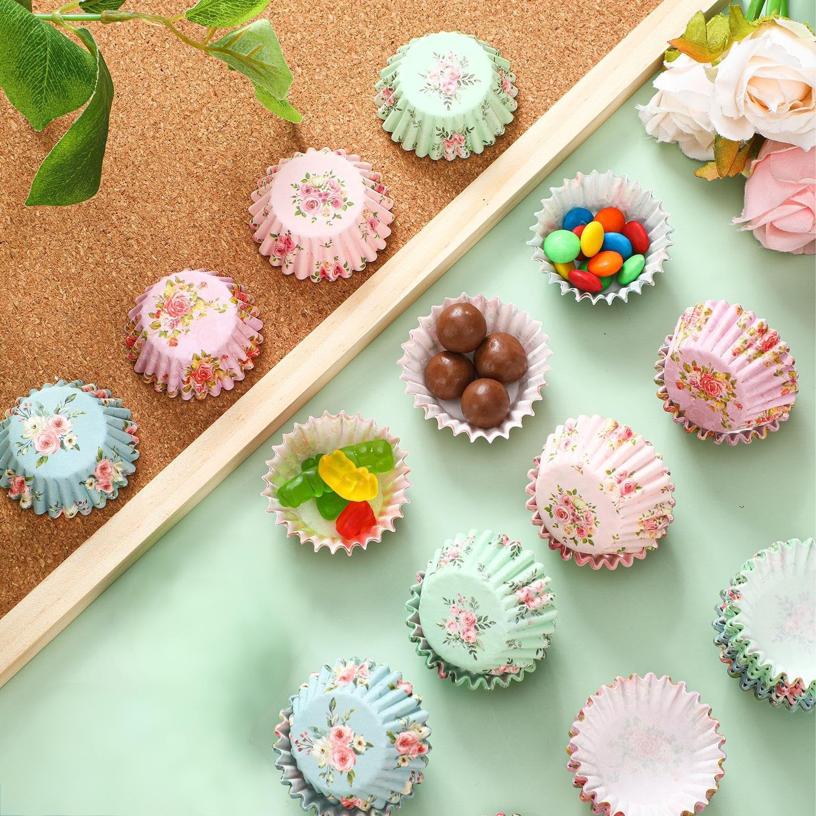 Assortment of floral-patterned mini cupcake liners filled with candies and chocolates on a light green surface with a cork background.