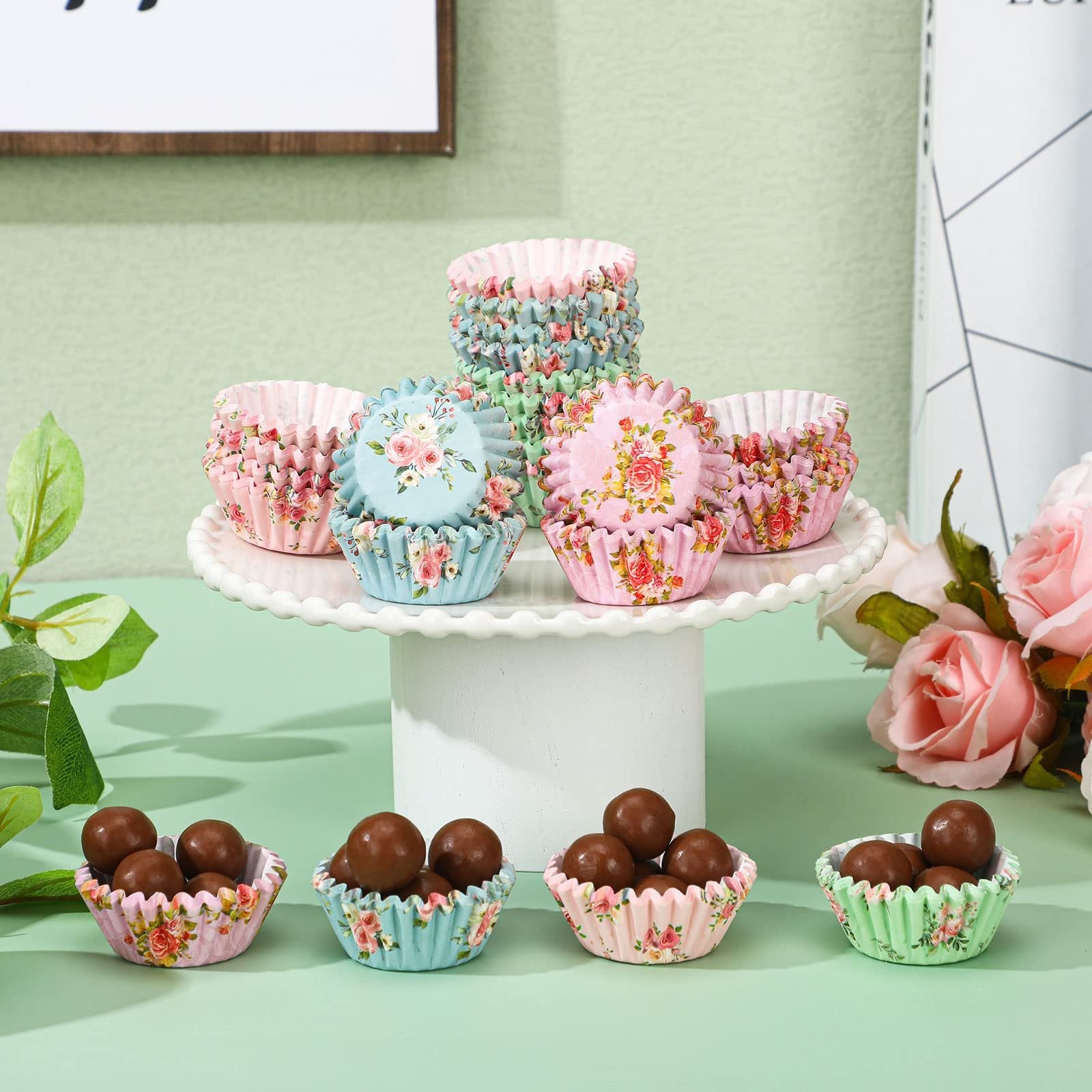 Assortment of floral print mini cupcake liners filled with chocolate candies, displayed on a white cake stand with decorative roses.