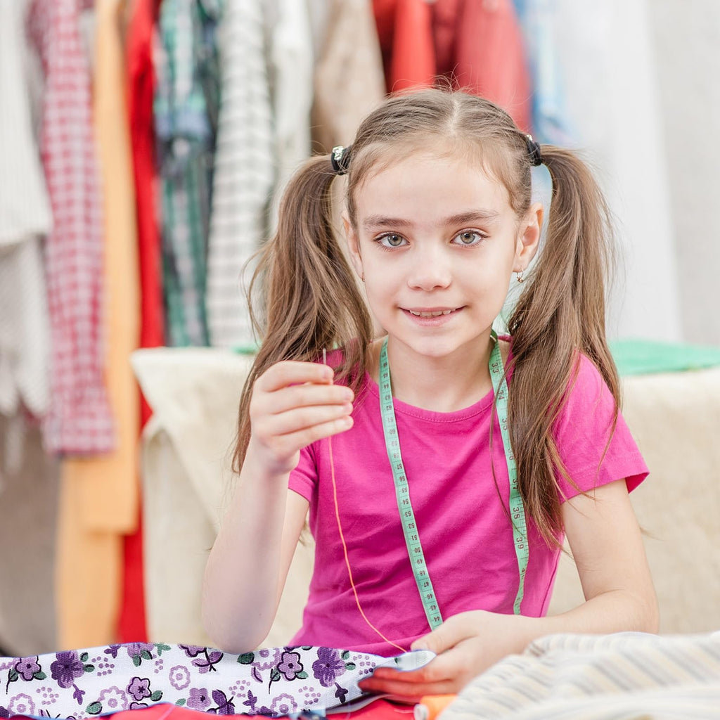 Young girl with pigtails sewing with a needle and thread, wearing a pink shirt and a green measuring tape around her neck.