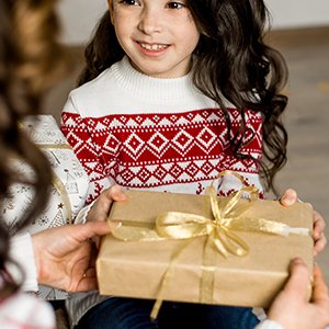 Young girl in a festive sweater receives a wrapped gift with a gold ribbon from an adult's hands.