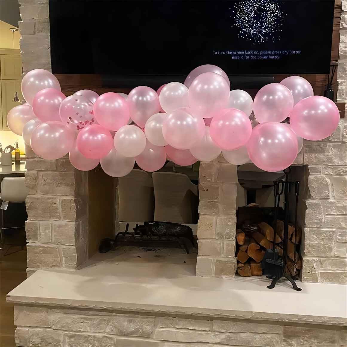 Light pink and white balloons draped over a stone fireplace mantel with a television screen in the background.
