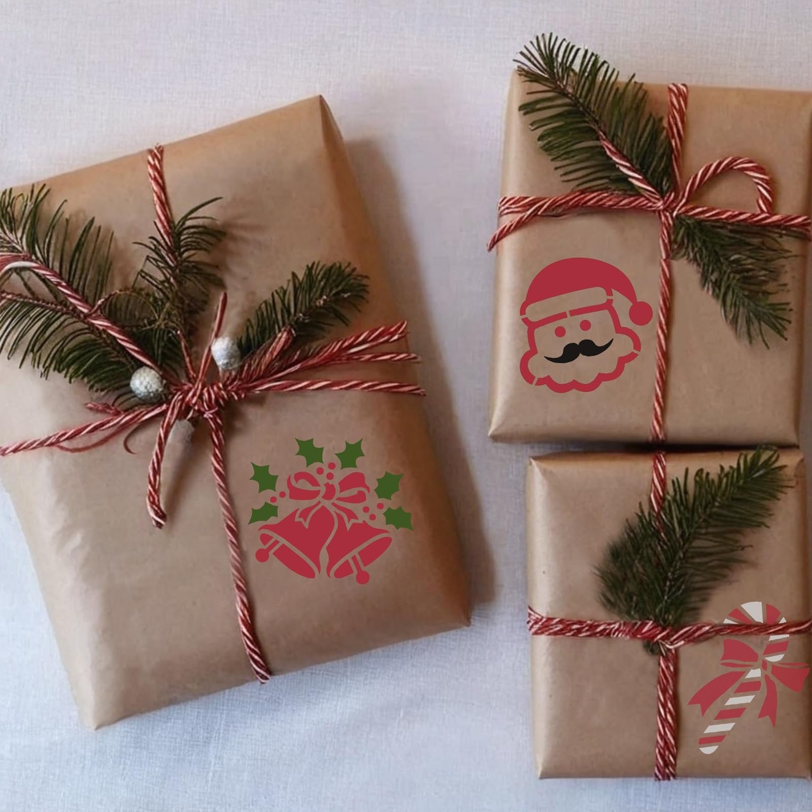 Three brown paper wrapped gifts decorated with pine sprigs and red and white twine, featuring Christmas stencils of Santa, bells, and a candy cane.