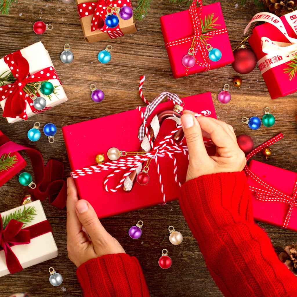 Hands wrapping a red gift box with colorful mini Christmas balls and pine sprigs scattered on a wooden table.