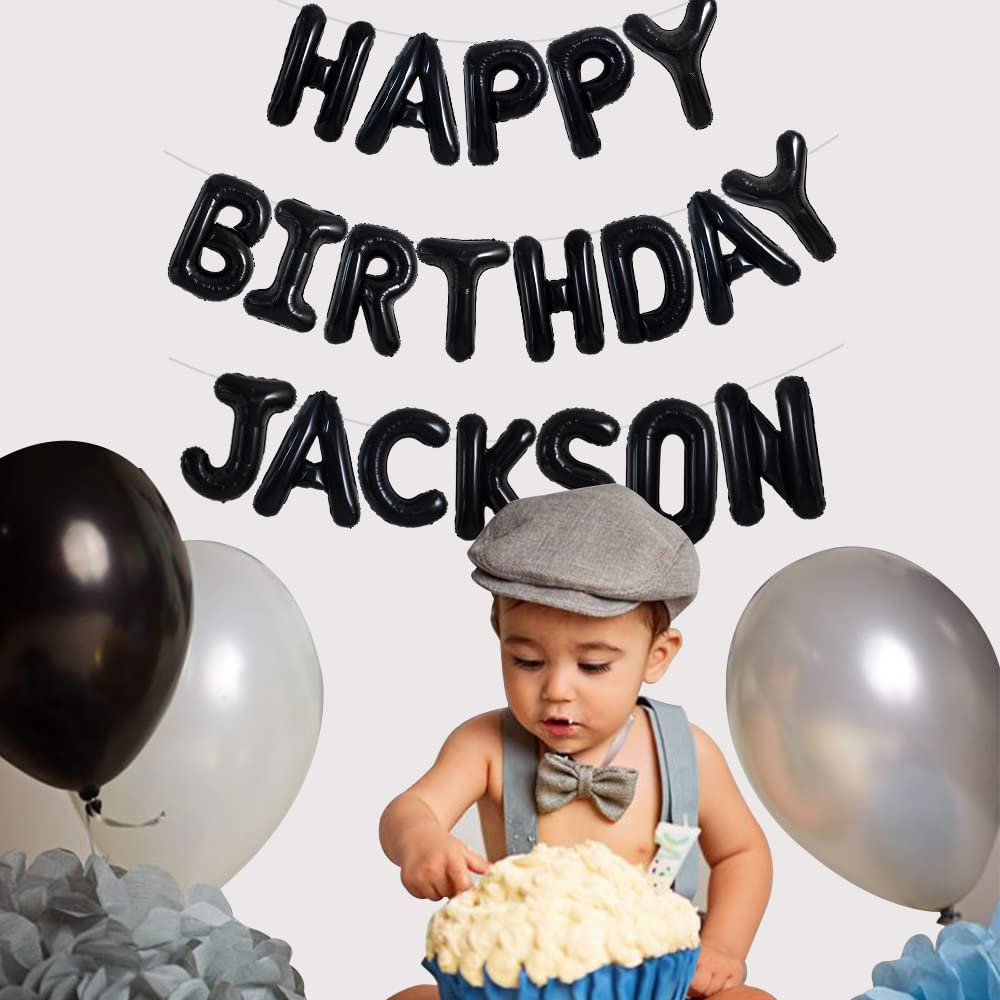 Baby boy in a cap and suspenders eating a giant cupcake with black 'Happy Birthday Jackson' balloons behind him.