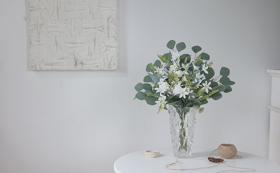 Artificial greenery stems with white flowers in a clear vase on a white table, with twine and a small ring nearby.