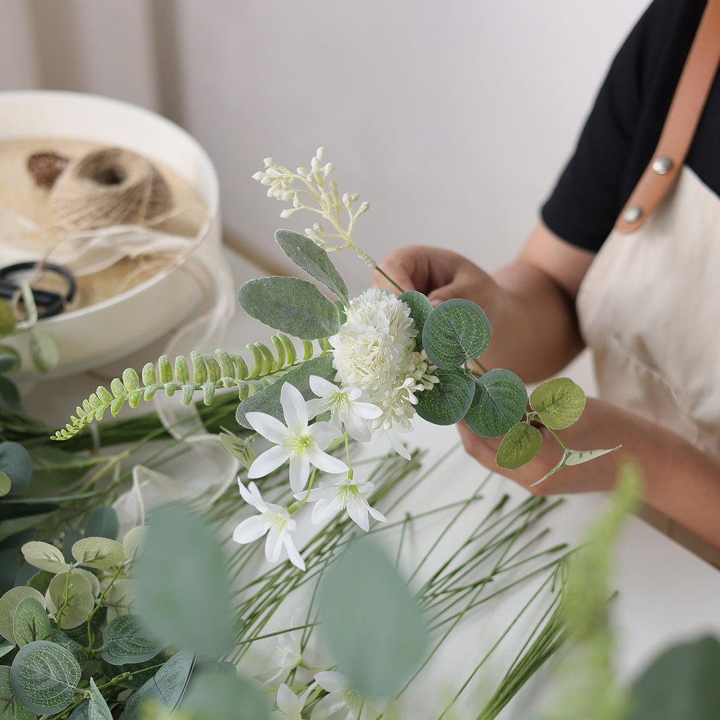 Hands arranging artificial white flowers and green eucalyptus stems on a white surface, with twine and scissors in a bowl nearby.