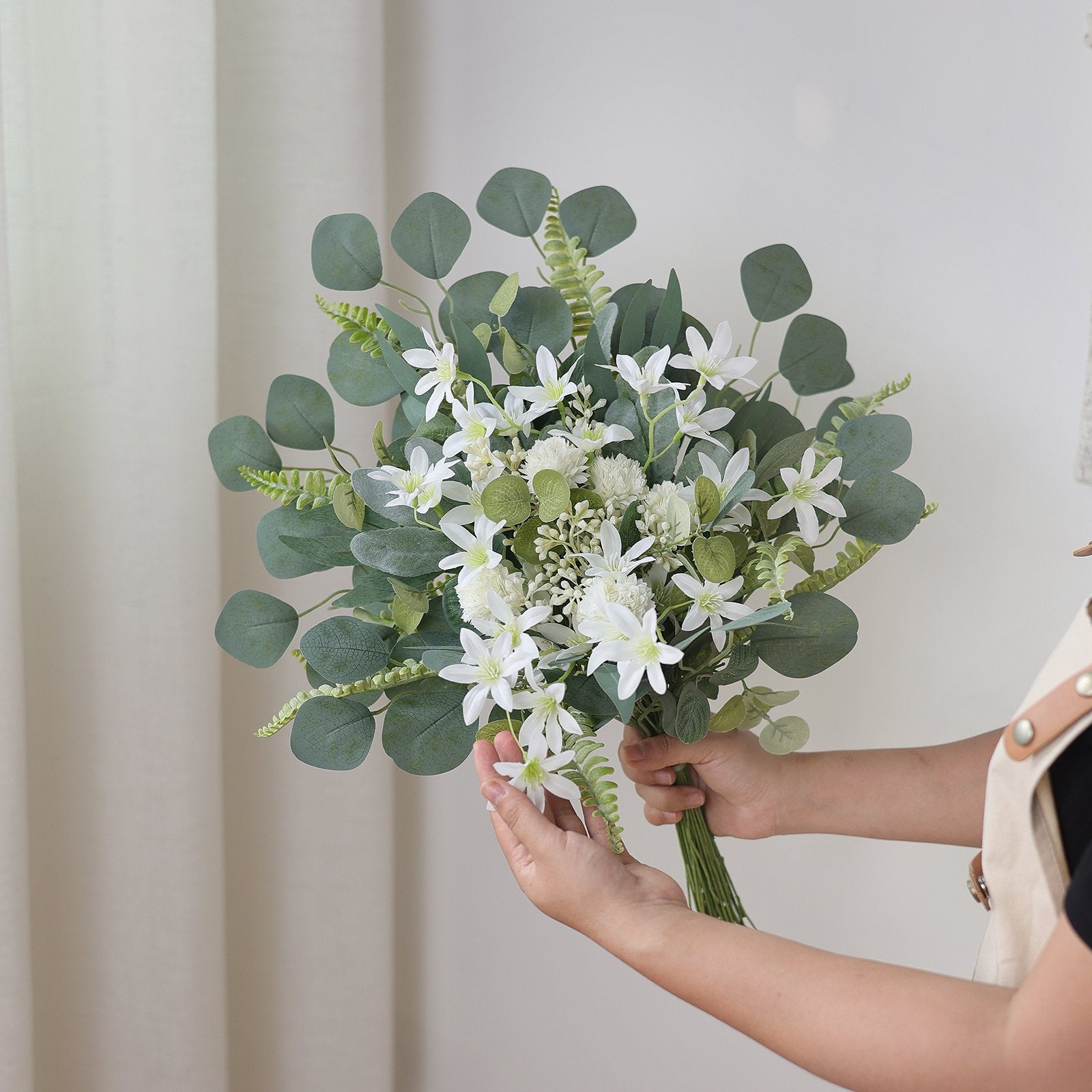 Hands arranging a bouquet of artificial eucalyptus leaves and white flowers against a neutral background.