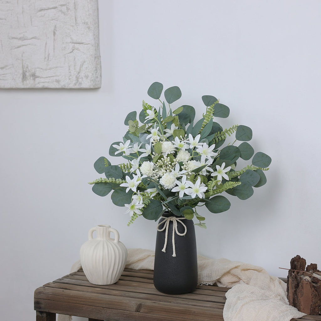Artificial greenery stems with white flowers in a black vase on a wooden table, next to a cream vase.