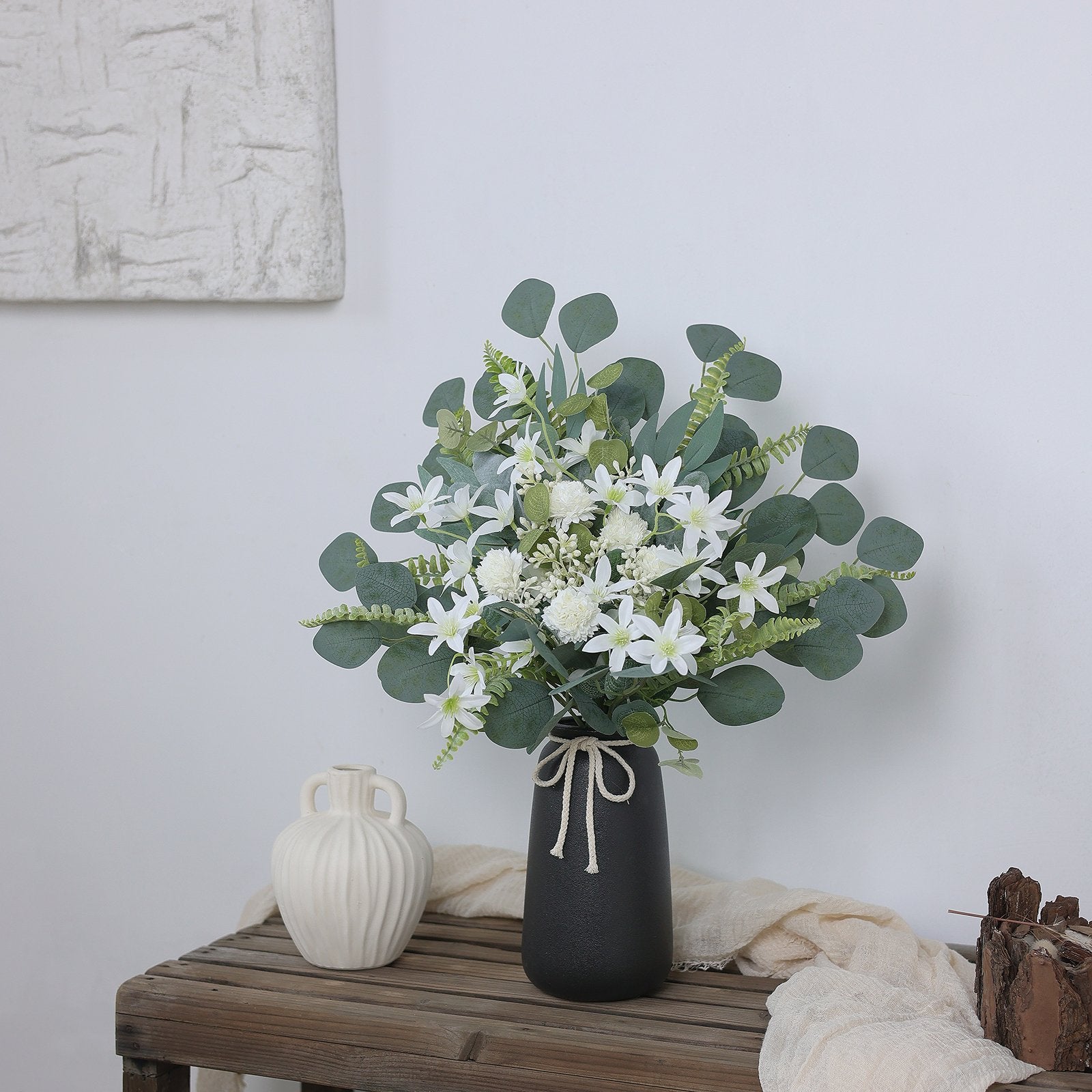 Artificial greenery stems with white flowers in a black vase on a wooden table, next to a cream vase.