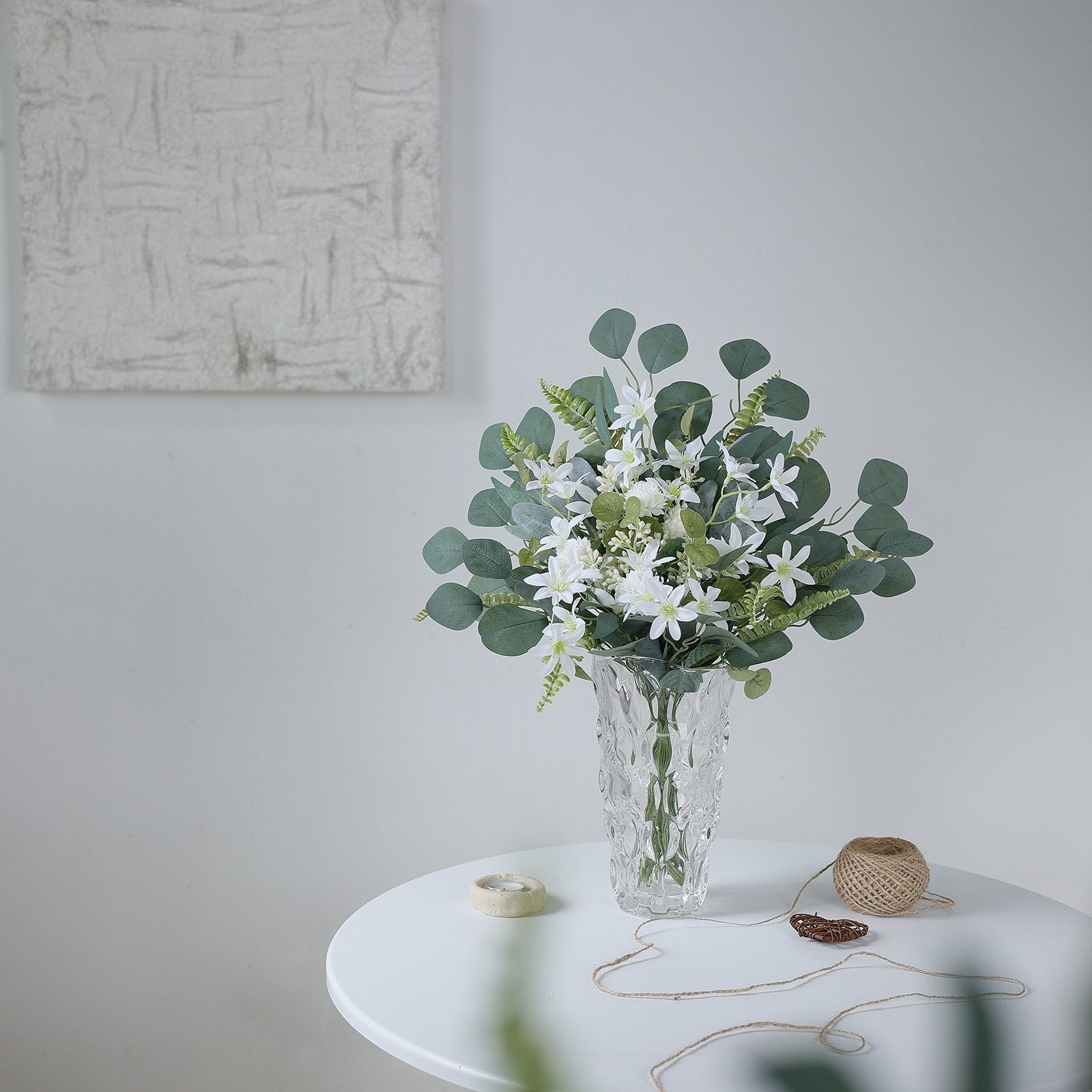 Artificial greenery stems with white flowers in a clear vase on a white table, with twine and a small heart decoration.