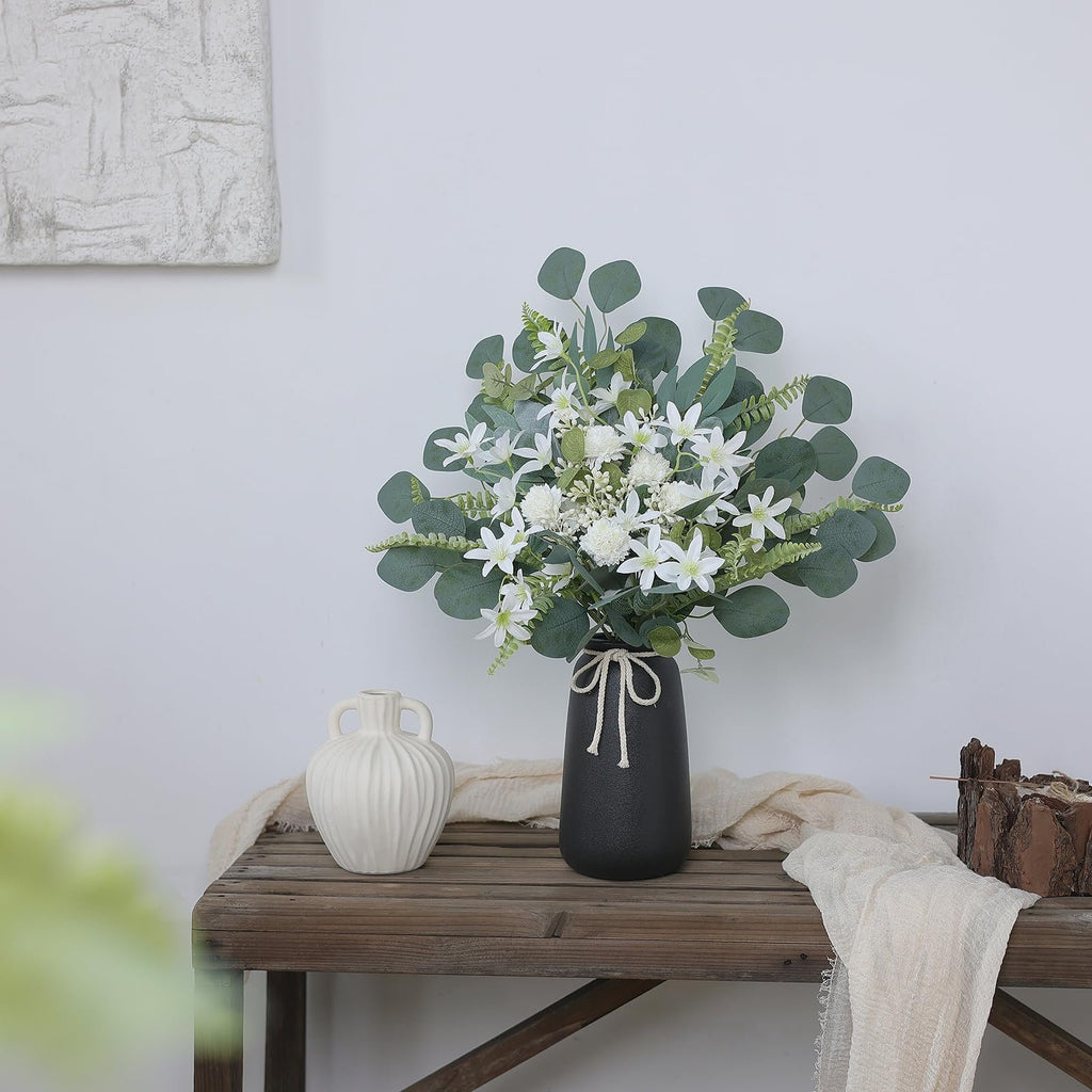Artificial greenery stems with white flowers in a black vase on a wooden shelf with a white vase and fabric.