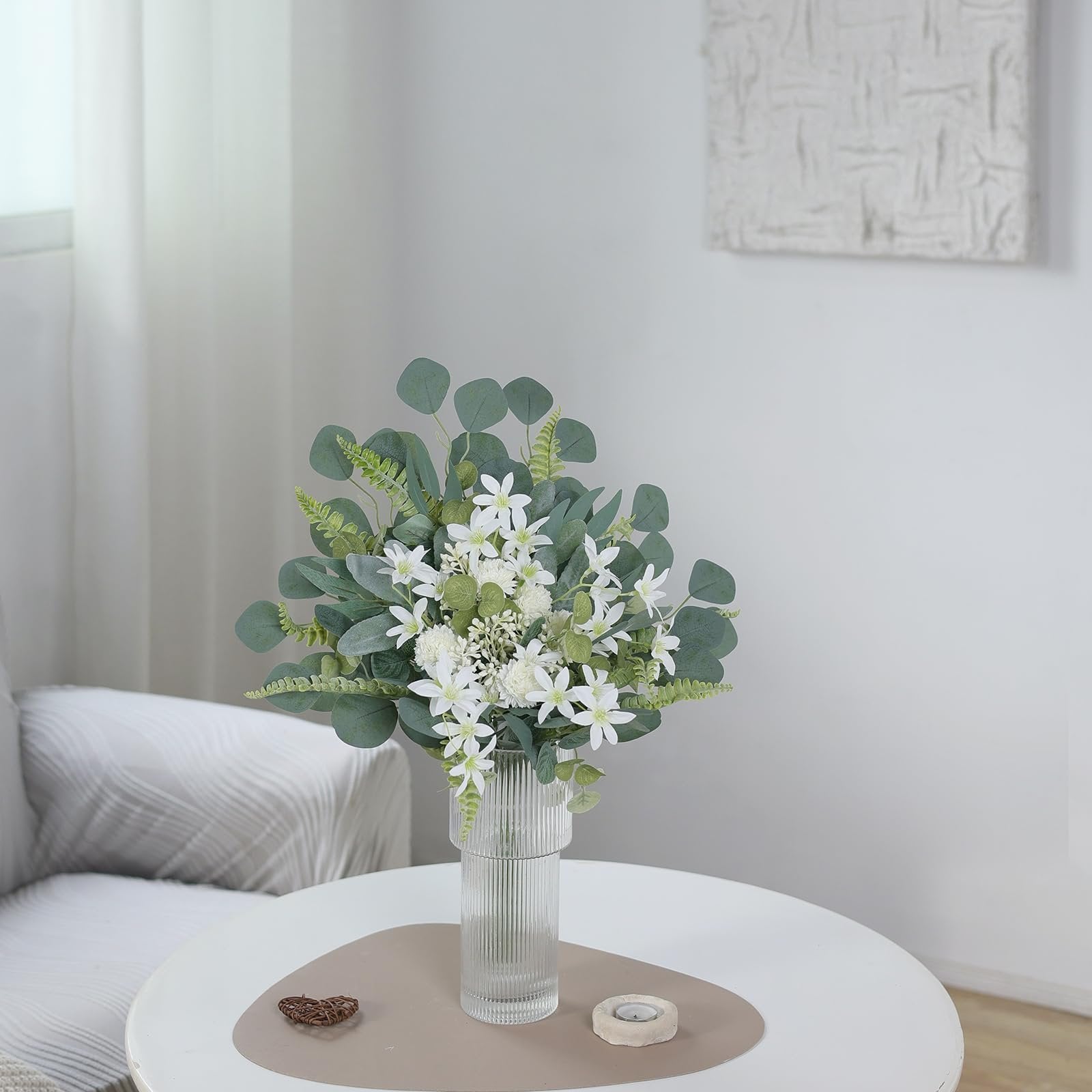 Artificial greenery bouquet with white flowers in a ribbed glass vase on a round table.