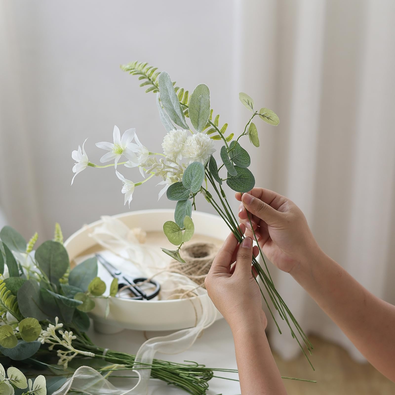 Hands arranging a bouquet of artificial white flowers and green leaves with twine and scissors in the background.