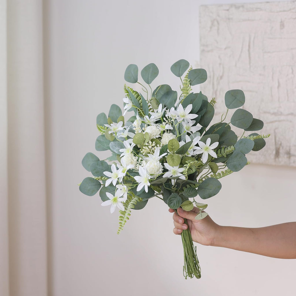 Hand holding a bouquet of artificial white flowers and green eucalyptus leaves against a textured wall.