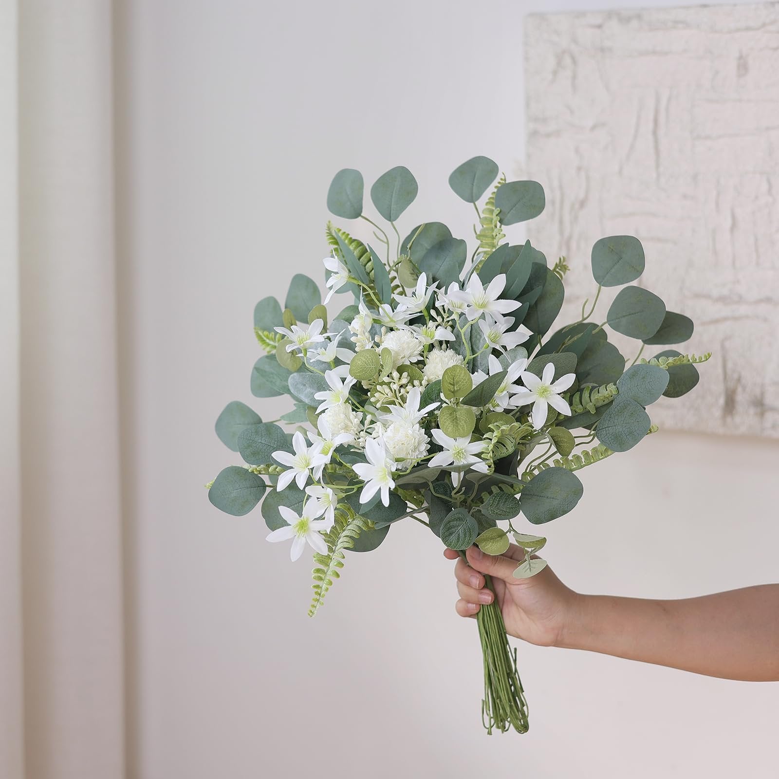 Hand holding a bouquet of artificial white flowers and green eucalyptus leaves against a textured wall.