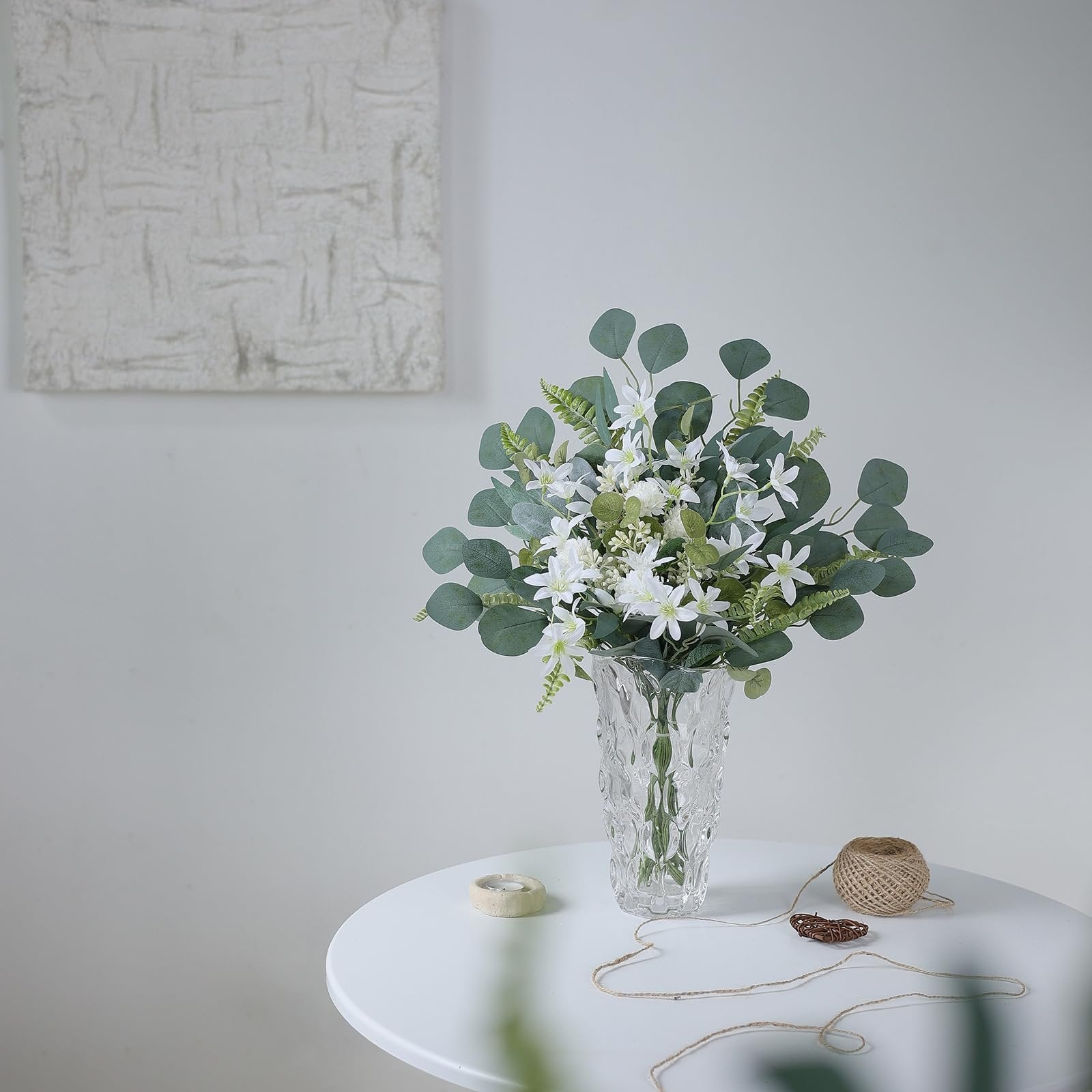 Artificial greenery bouquet with white flowers in a crystal vase on a white table, with twine and a small candle nearby.