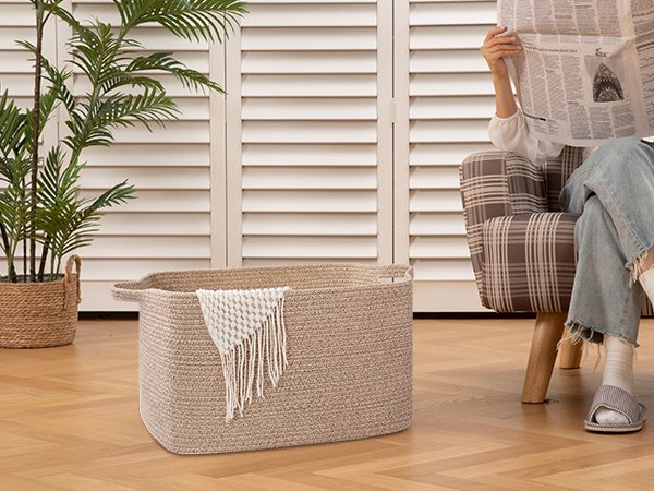 Large woven storage basket with handles, holding a folded blanket, next to a chair where a person reads a newspaper.