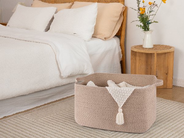 Large beige cotton rope basket with handles filled with a folded white blanket, placed on a rug next to a bed.