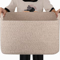 Person holding a large beige woven cotton rope storage basket with handles, front view.