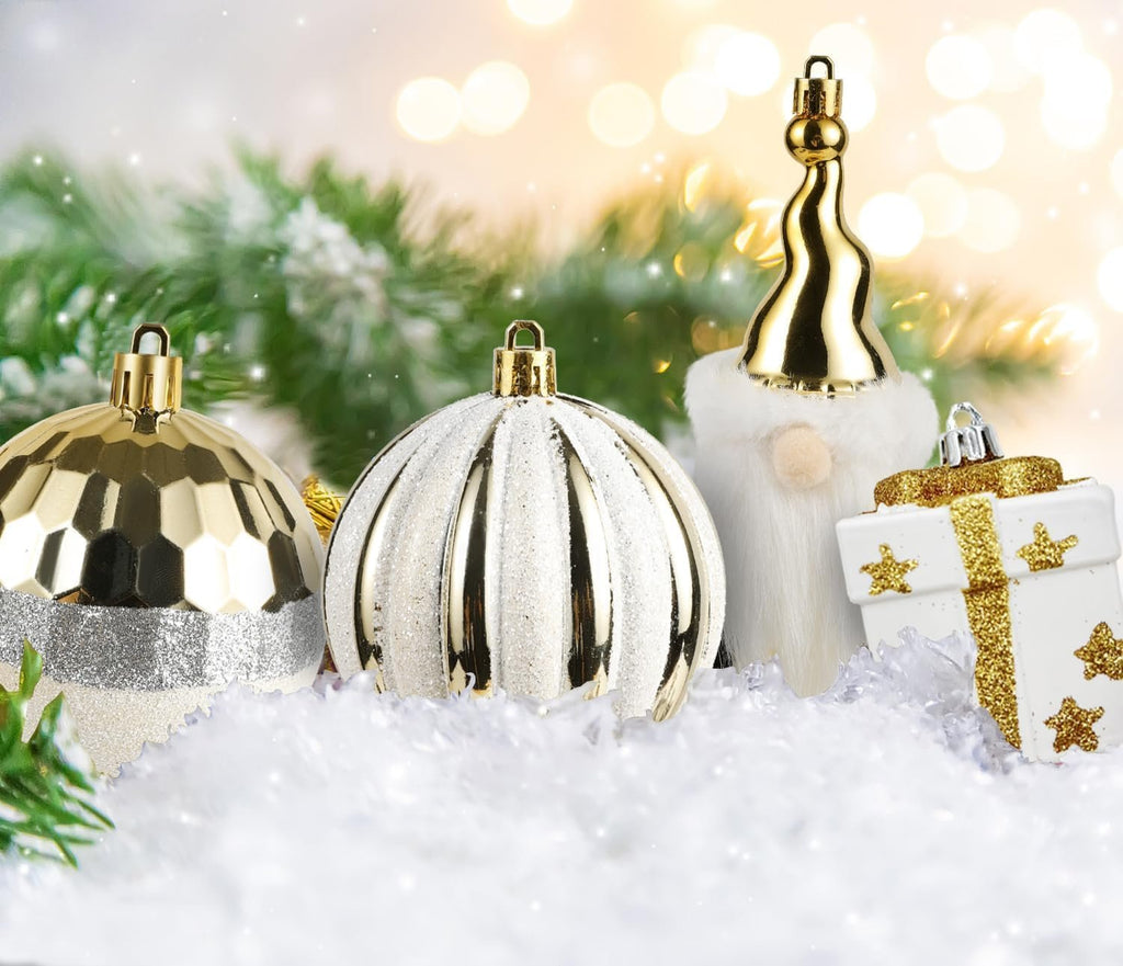 Assorted white and gold Christmas ornaments including a faceted ball, a ribbed ball, a gnome figure, and a gift box shape, arranged on faux snow with pine branches and bokeh lights.