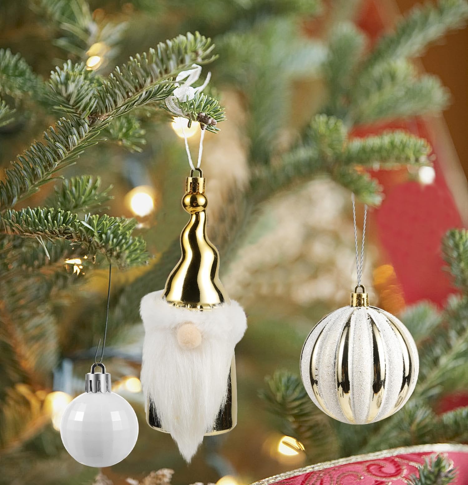 Three Christmas ornaments hanging on a tree: a white ball, a gold and white gnome, and a gold and white ribbed ball.