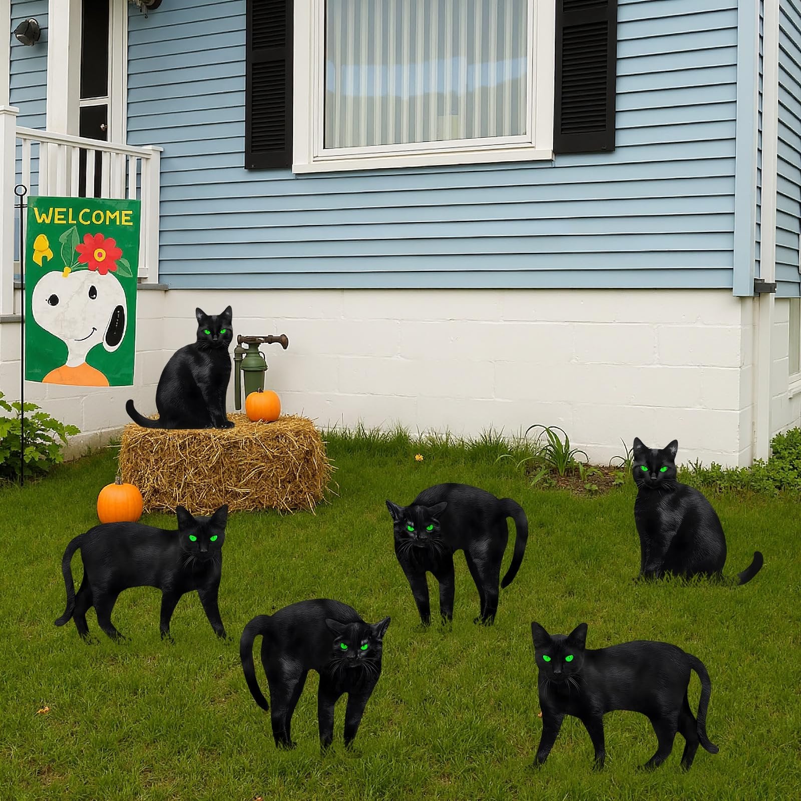 Six black cat silhouette yard signs with green glowing eyes arranged on a lawn in front of a blue house, with a welcome flag and hay bale.