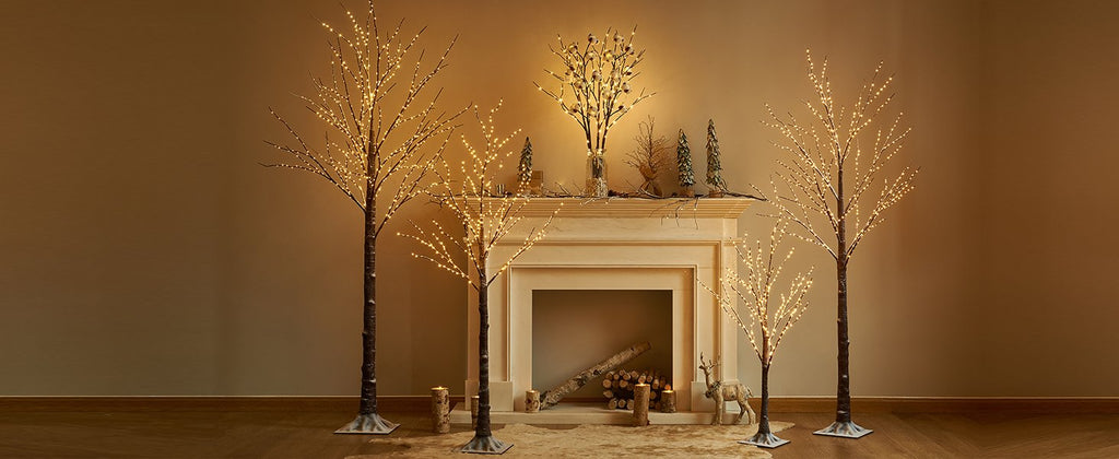 Four illuminated twig trees and a vase of lighted branches arranged around a fireplace with decorative elements.