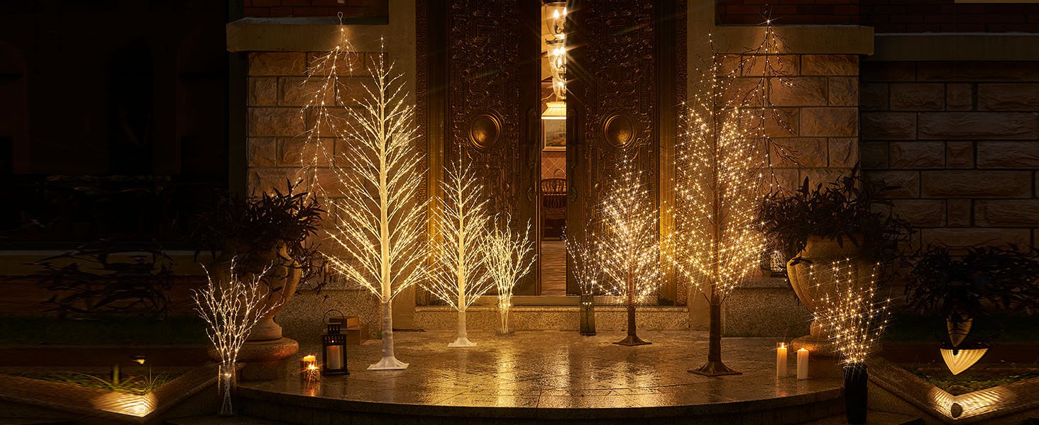 Several illuminated twig trees and branches with fairy lights displayed outdoors at night in front of a large ornate door.