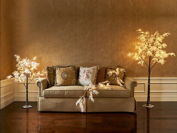 Two lighted twig trees with white leaves and fairy lights flank a sofa with decorative pillows and a garland.