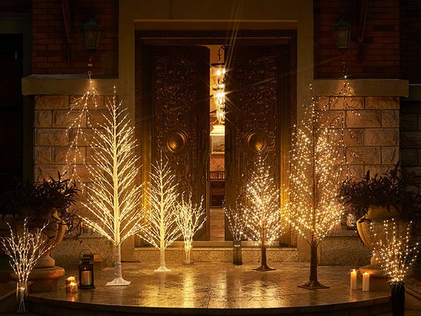 Group of illuminated twig trees and branches with fairy lights displayed on a porch at night.