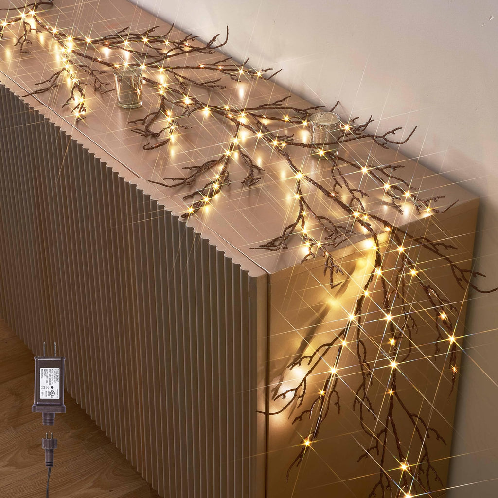 Brown twig garland with 120 warm white fairy lights draped on a ribbed cabinet, with a plug-in adapter visible.