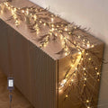 Brown twig garland with 120 warm white fairy lights draped on a ribbed cabinet, with a plug-in adapter visible.