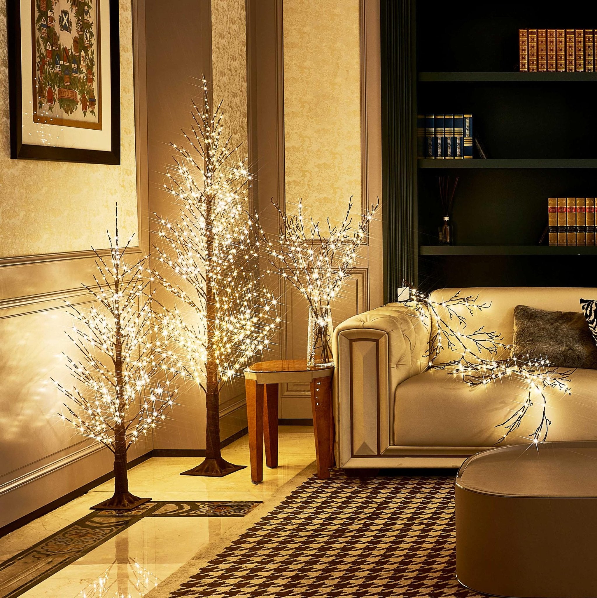 Three illuminated twig trees and a lighted branch garland arranged in a living room setting with a sofa and bookshelf.