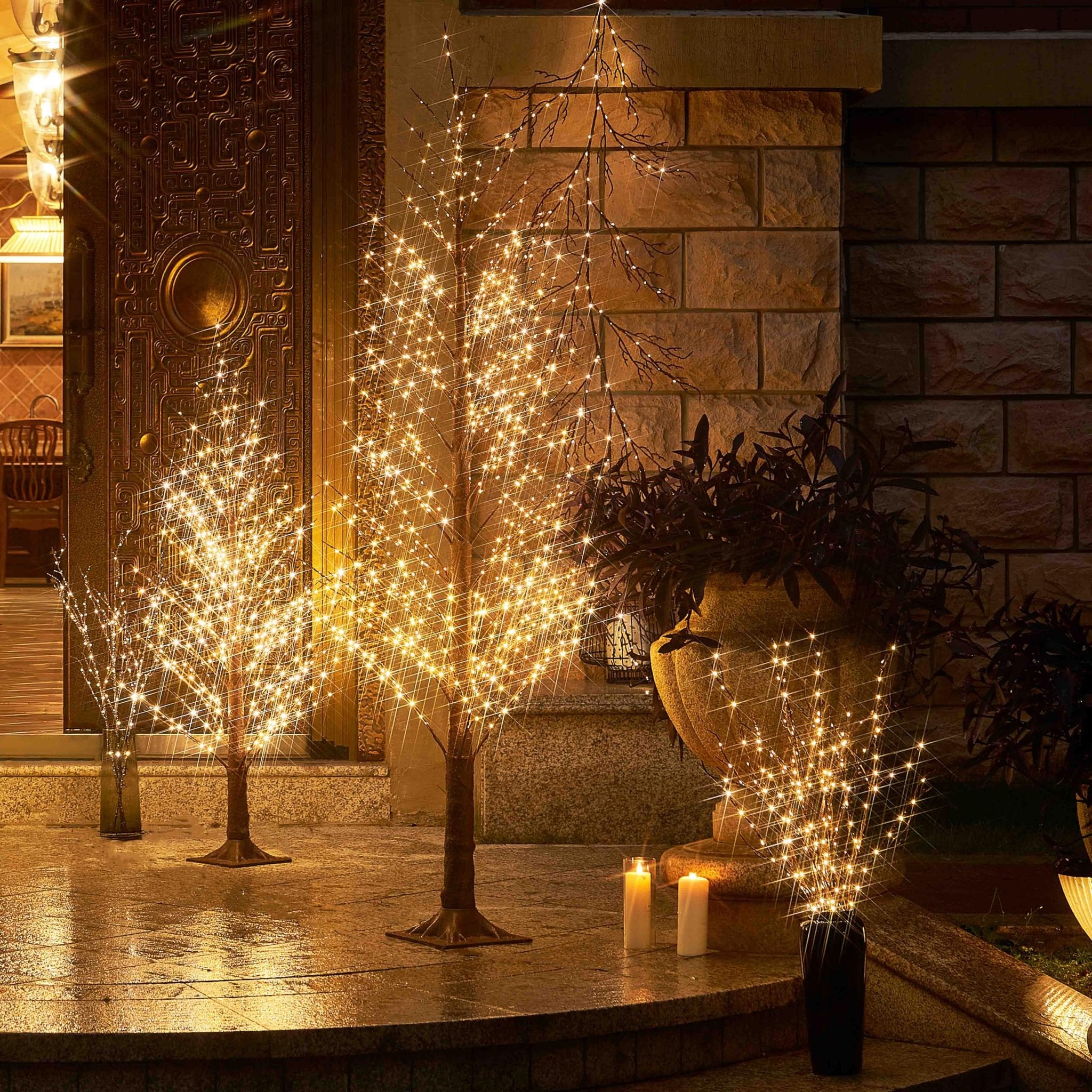Two illuminated twig trees and a vase of lights on a wet stone patio at night, with a decorative door and wall in the background.