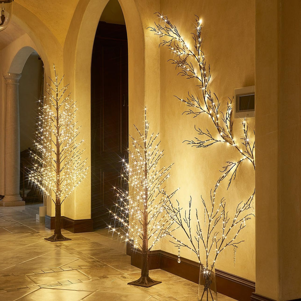 Three lighted twig trees and a wall-mounted twig garland with warm white fairy lights in an indoor hallway.